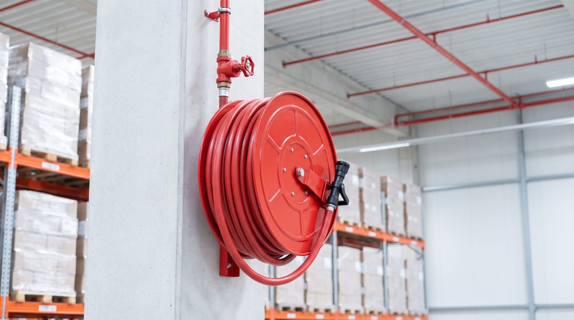 Robinet d'incendie armé fixé sur un poteau en béton d'un entrepôt neuf, dévidoir rouge avec son tuyau enroulé et sa lance, vanne rouge sur l'alimentation, rayonnages de palettes au fond