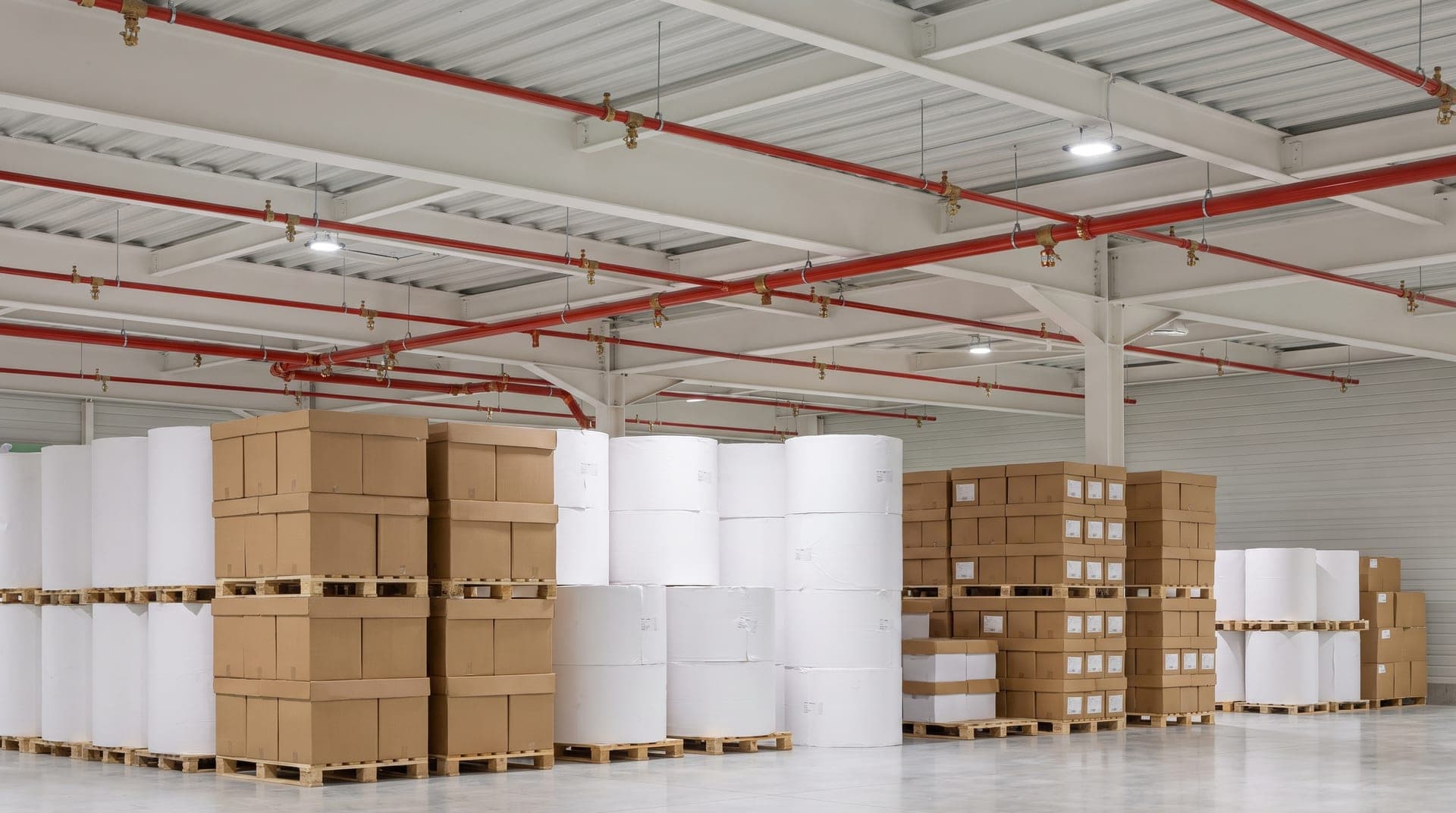 Magasin de produits finis d'une papeterie, grosses bobines de papier blanc et cartons sur palettes, réseau sprinkler rouge et têtes en laiton sous la charpente