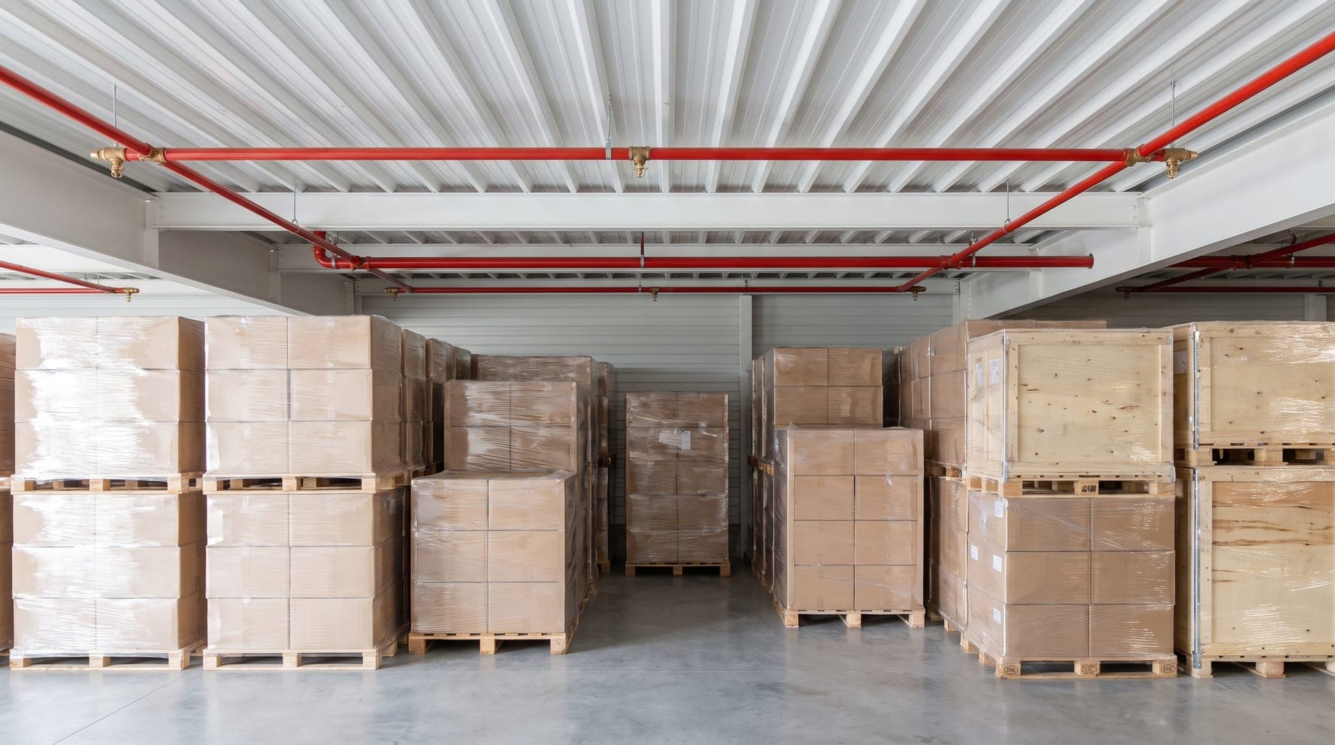 Entrepôt agroalimentaire, palettes de cartons filmés et caisses en bois, canalisations sprinkler rouges et têtes en laiton sous le bac acier