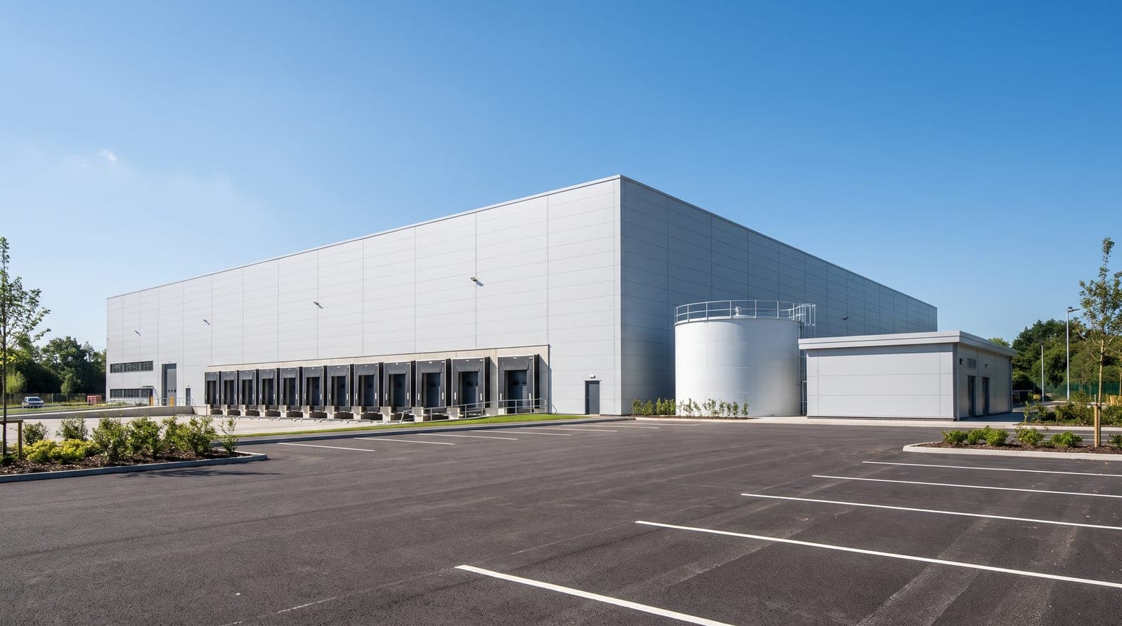 Plateforme logistique neuve en bardage gris clair vue de l'extérieur par beau temps, quais de chargement alignés, cuve sprinkler cylindrique et petit local technique sur le côté, parking vide et propre