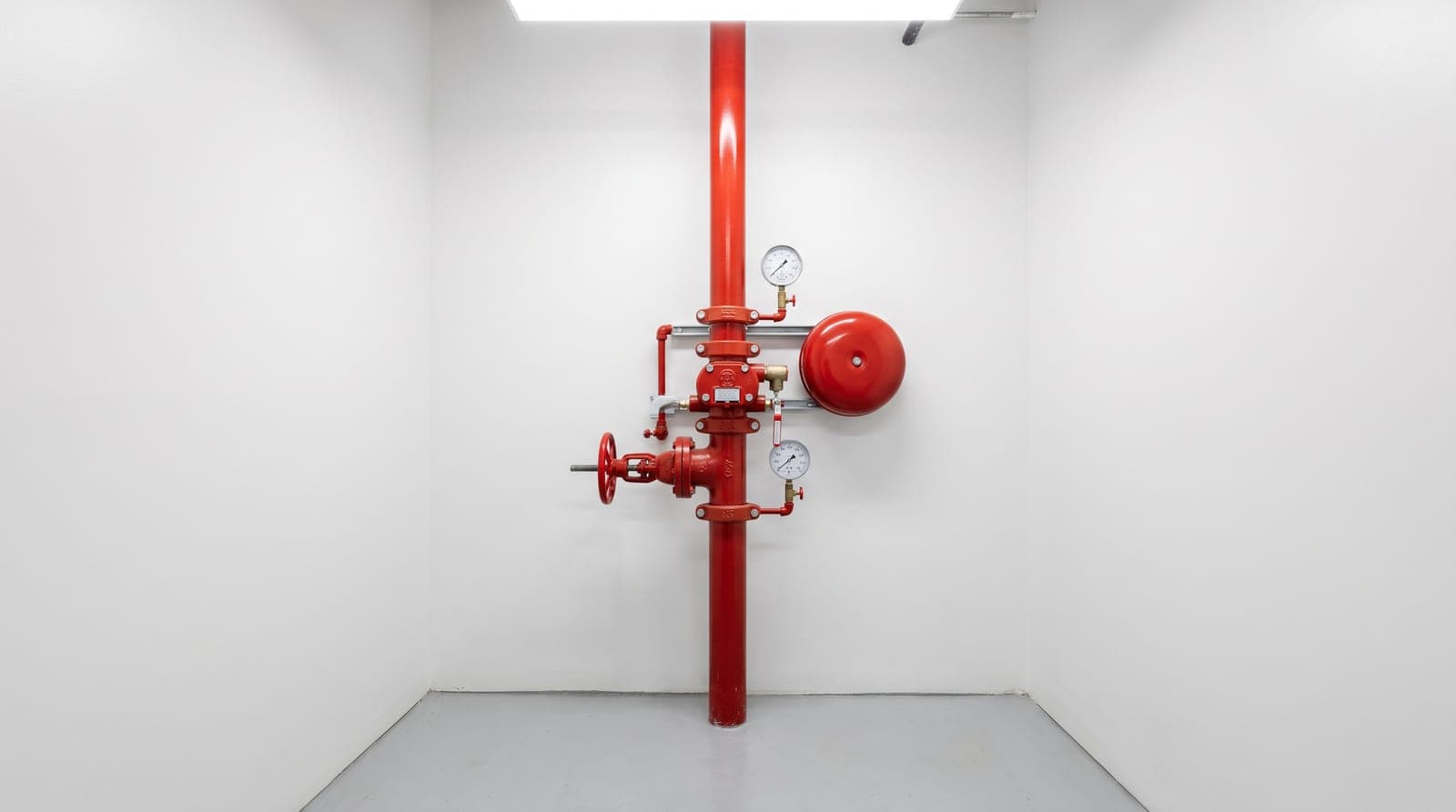 Poste de contrôle sprinkler neuf dans un local technique propre aux murs blancs : colonne de canalisations rouges, vanne d'arrêt, clapet d'alarme, deux manomètres et gong hydraulique, sol peint gris, éclairage net