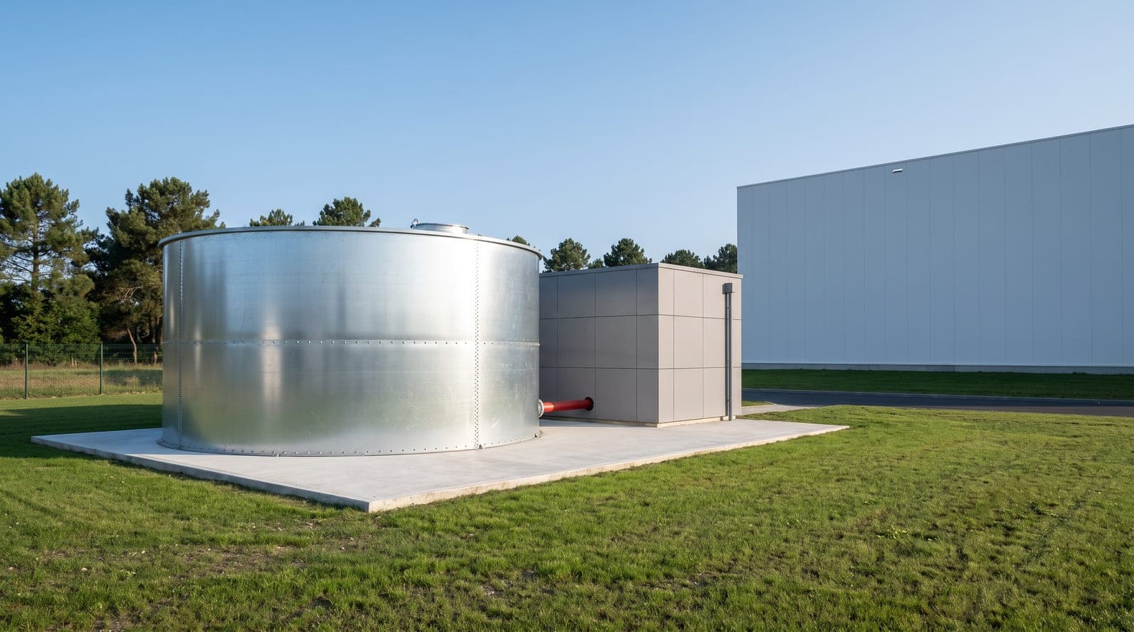 Cuve sprinkler cylindrique en acier galvanisé neuve, posée sur dalle béton à côté d'un petit bâtiment technique en bardage gris, pelouse tondue et ciel clair, arrière-plan d'entrepôt moderne