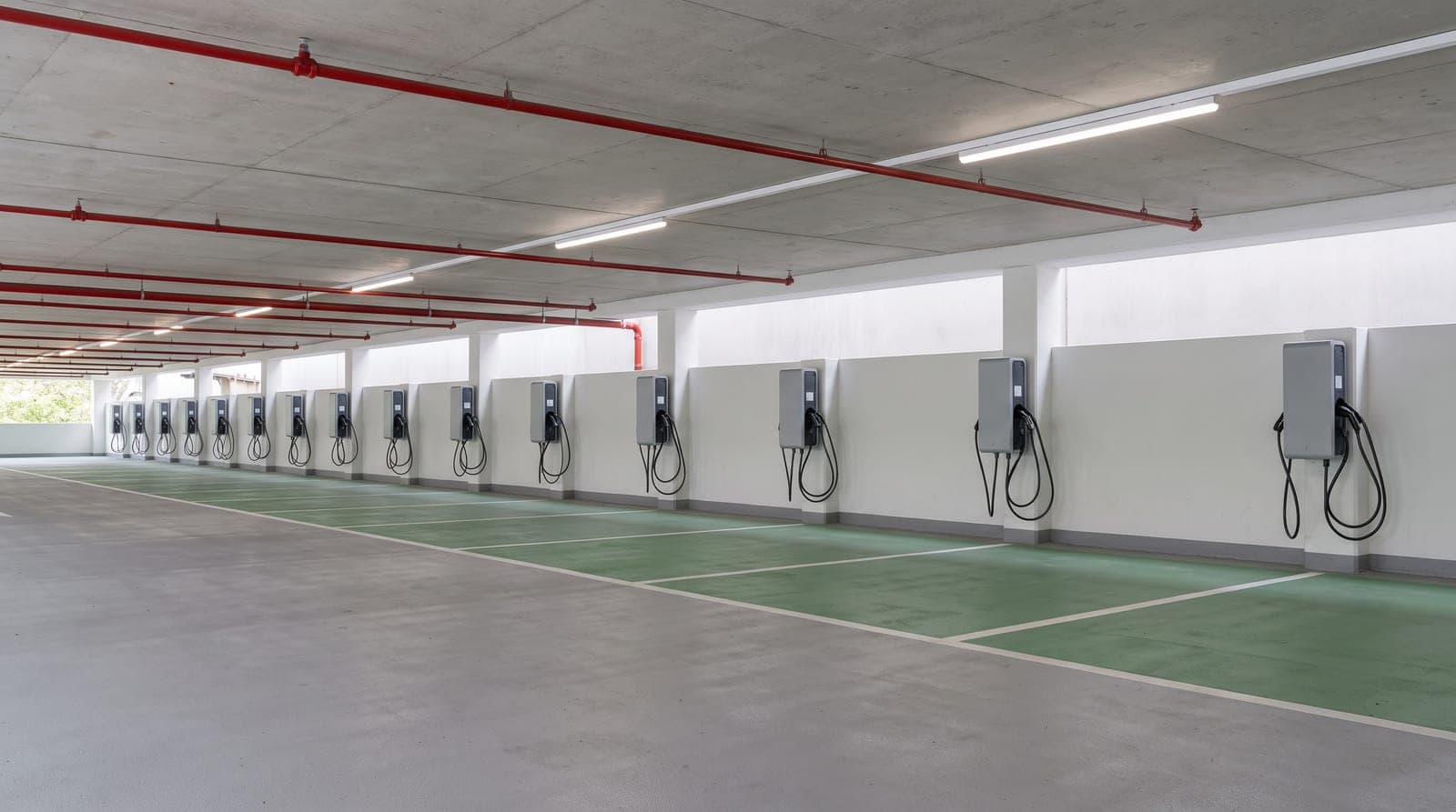 Rangée de bornes de recharge murales neuves dans un parking couvert propre, places vides au sol gris et marquage vert, réseau sprinkler rouge au plafond au-dessus des bornes, lumière blanche