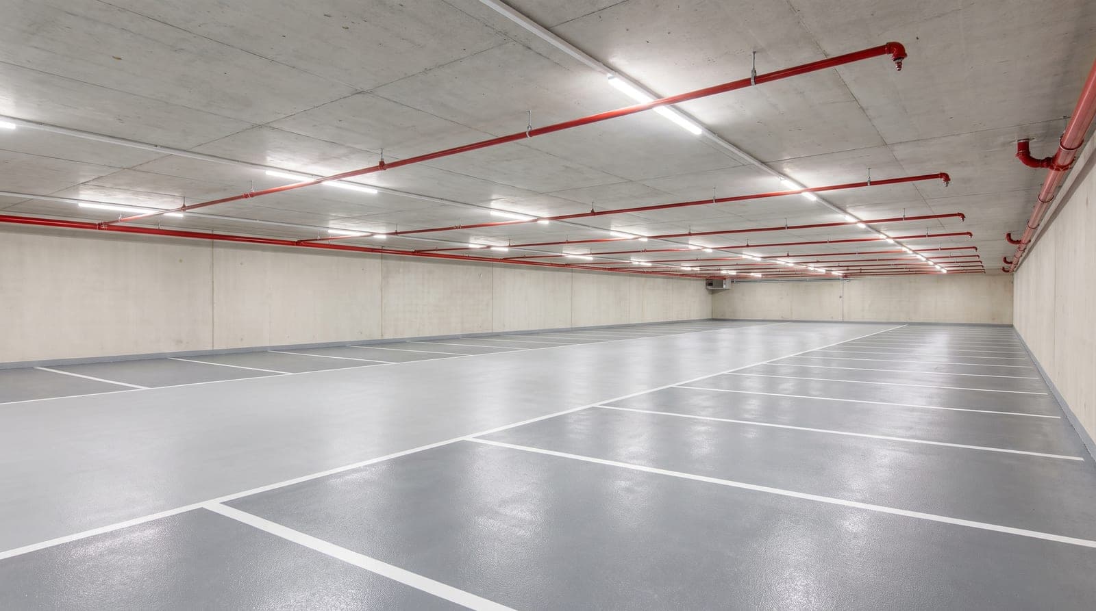 Niveau de parking souterrain neuf et vide, sol résine gris avec marquage blanc net, plafond béton clair équipé d'un réseau sprinkler rouge et de têtes régulières, éclairage LED uniforme