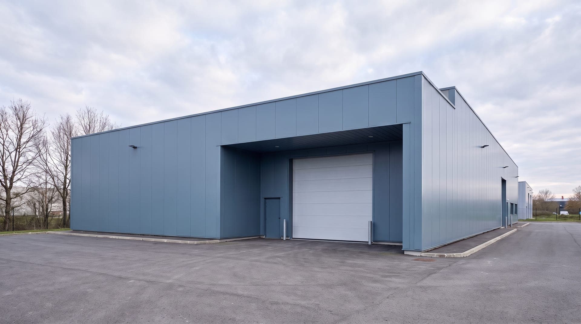 Bâtiment industriel neuf en bardage bleu gris, grande porte sectionnelle sous auvent, voirie enrobée et arbres sans feuilles