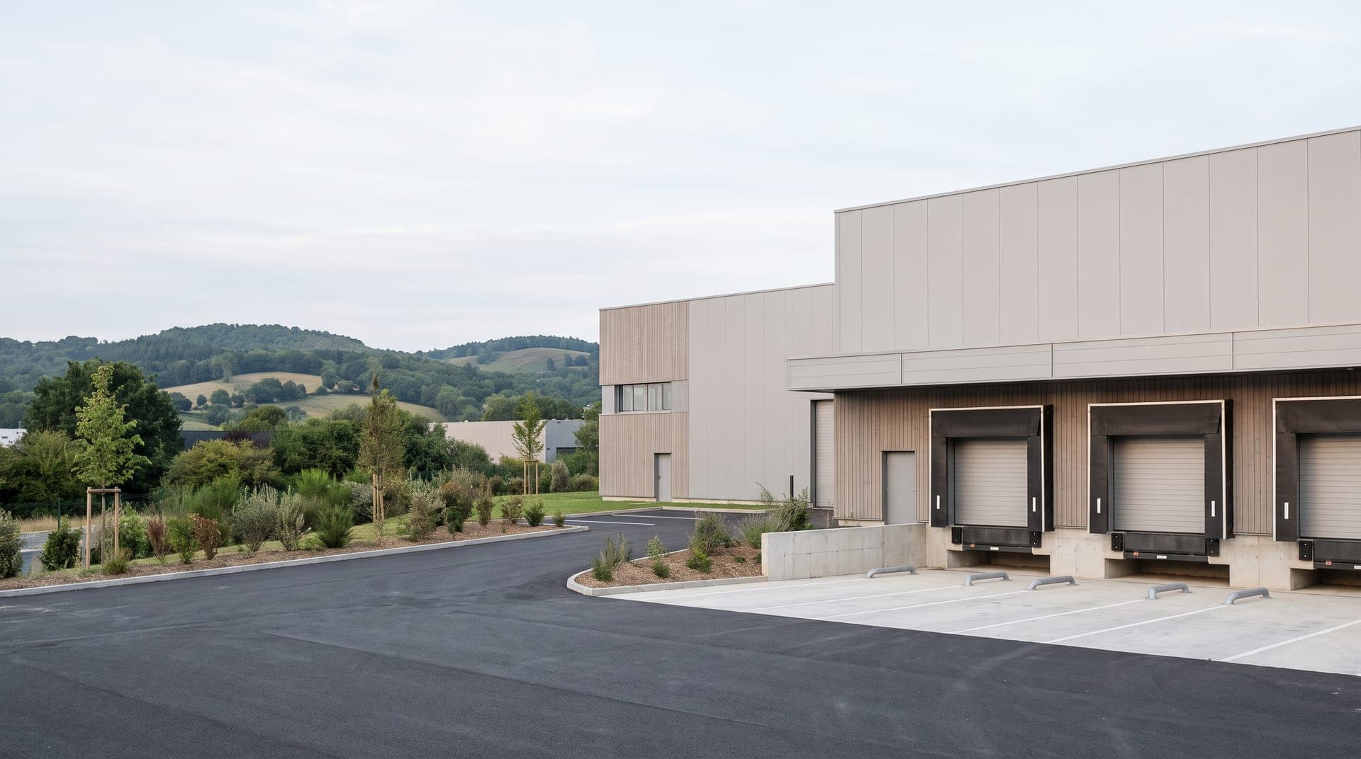 Bâtiment industriel neuf en bardage clair, trois quais de livraison, voirie neuve et collines boisées au fond