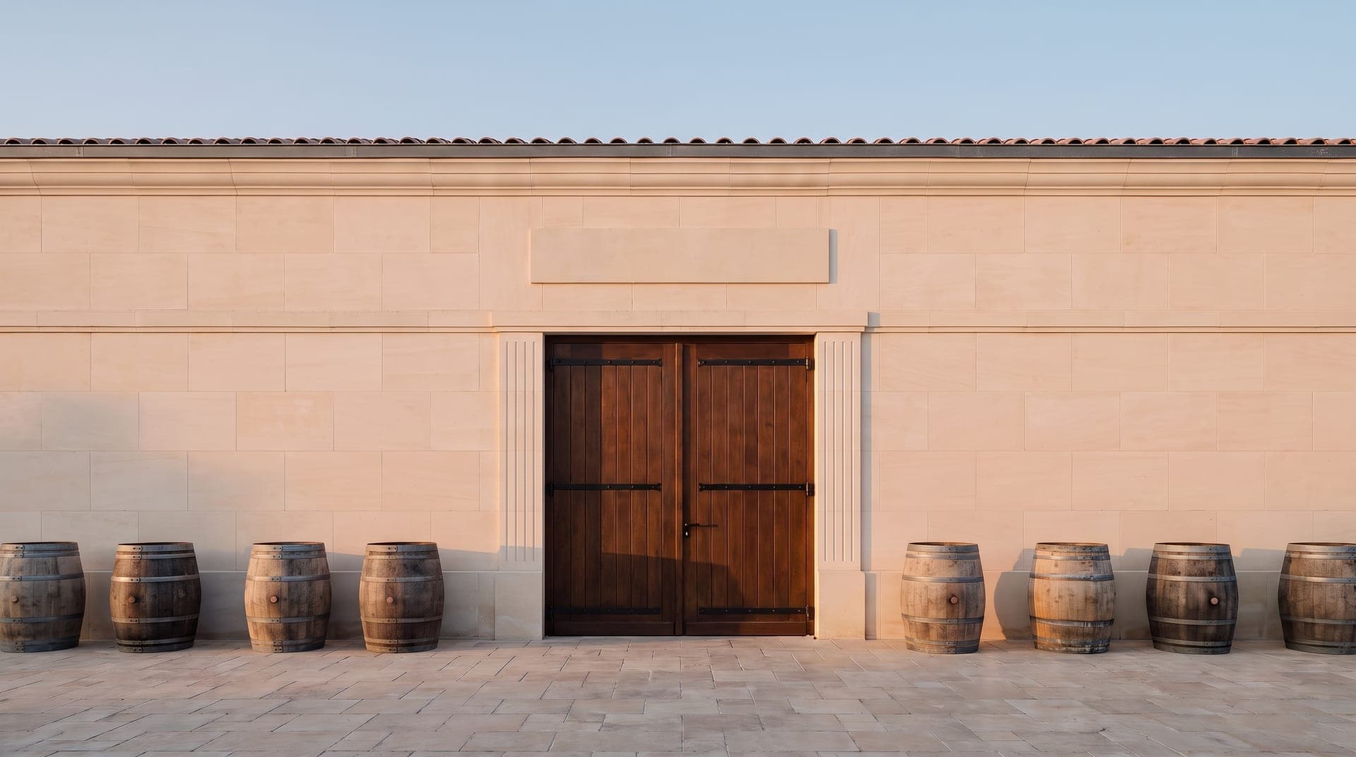 Façade en pierre claire d'un chai, grande porte en bois, fûts de chêne alignés devant le mur
